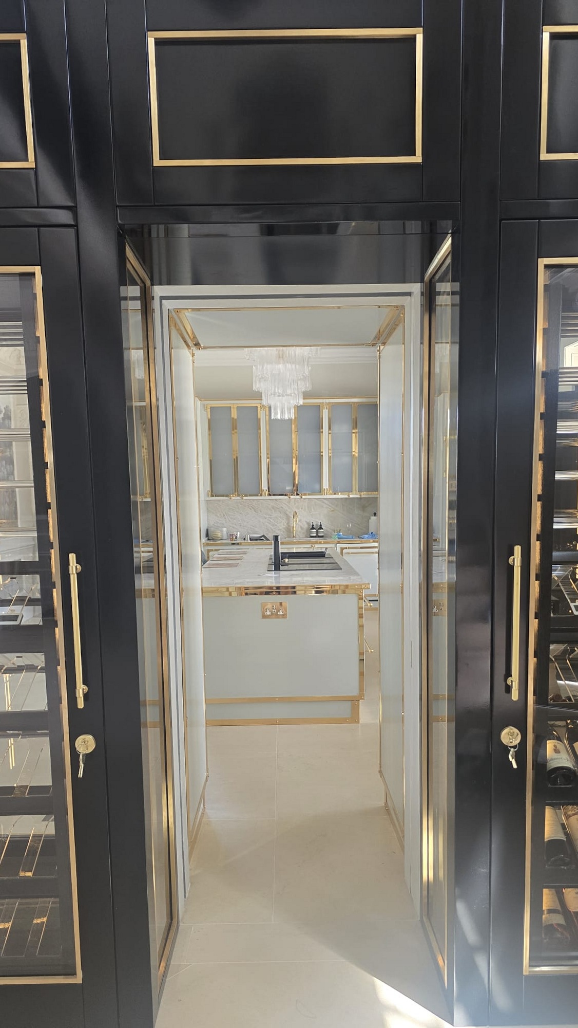 Looking from the butler's pantry into the kitchen of a Cotswolds country house, with brass-trimmed cabinetry and glass-fronted wine display
