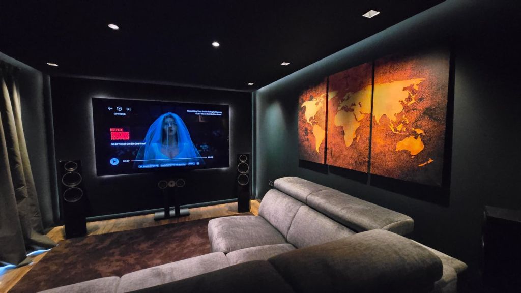 Garden room cinema interior — wide view showing the off-black ceiling with coffee-table downlighters, world-map feature artwork, grey sectional seating and the bias-lit Sony 85