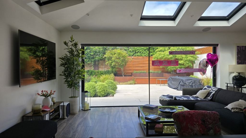 Open-plan living space with wall-mounted TV and in-ceiling speakers opening onto a landscaped garden