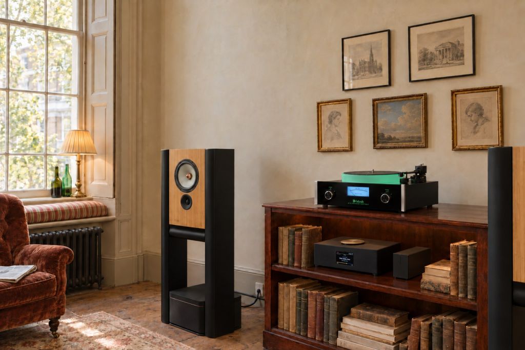 Illustration of a Grimm Audio LS1c loudspeaker beside a low mahogany bookcase carrying a McIntosh MT10 turntable, with a Grimm MU1 streamer and PW1 phono stage on its shelf among old books, in a Georgian book room with a shuttered sash window, a ticking-covered window seat and a group of small framed prints.